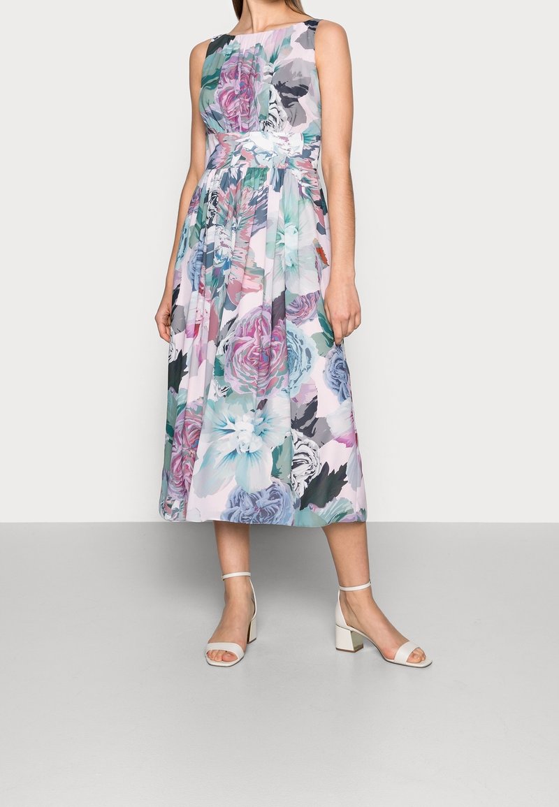 Femme portant une robe fleurie sans manches, longueur mi-cuisse, avec des fleurs roses, bleues et vertes, associée à des sandales blanches à talons carrés et ouvertures à l'avant.