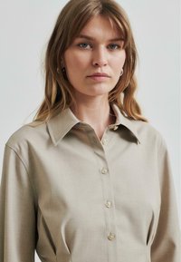 Beige skjorte med krage, knappestClosure og lange ermer, med en glatt tekstur og subtil glans. Nærbilde av øverste halvdel.