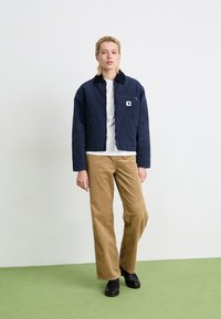 Person wearing a navy blue jacket, white shirt, tan corduroy pants, and black shoes standing on green floor against white wall.