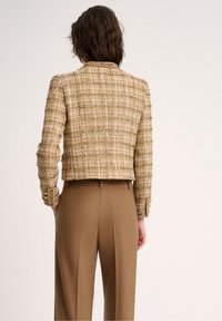 Texturovaná zkrácená bunda z metalického hnědo-zlatého tweedu s límcem, detailními knoflíky a spárovaná s hnědými kalhotami s vysokým pasem.