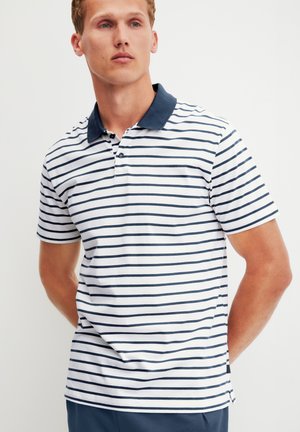 Mann trägt weißes Poloshirt mit marineblauen horizontalen Streifen und einfarbigem marineblauen Kragen, steht mit den Händen hinter dem Rücken vor einfarbigem Hintergrund.