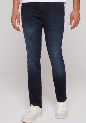 Dark blue denim jeans with a slim fit, featuring five pockets and subtle whiskering. Paired with white sneakers for a casual look.