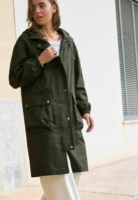 Impermeabile verde oliva con cappuccio, dotato di bottoni a pressione, polsini elasticizzati, due tasche frontali e vestibilità comoda, indossato sopra una maglietta bianca.