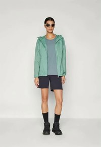 Kvinna som bär solglasögon, grön huva jacka, blå skjorta, svarta shorts, svarta strumpor till mitten av vadarna och svarta vandringsskor som står framför en enfärgad bakgrund.