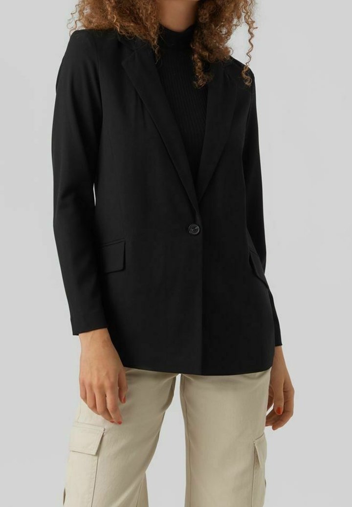 Femme portant un blazer noir à un seul bouton sur un haut noir, associée à un pantalon cargo beige, debout devant un fond clair uni.