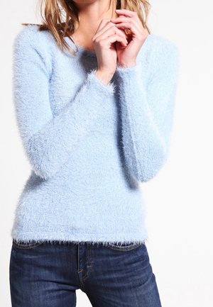 Kobieta w jasnoniebieskim, puszystym swetrze z długim rękawem i ciemnoniebieskich dżinsach, z delikatnie złączonymi rękami przy twarzy na białym tle.