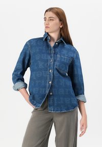 Modell med denimskjorte med subtilt tekstmønster, kombinert med lyse bukser, poserer selvsikkert mot en enkel hvit bakgrunn.