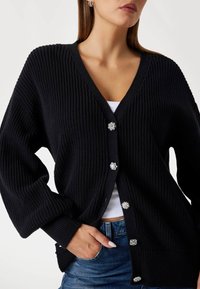 Black ribbed cardigan featuring a V-neck, oversized sleeves, and decorative buttons with pearl and metallic accents. Textured knit material.