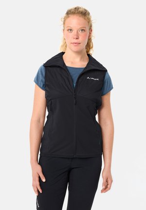 Junge Frau trägt eine schwarze ärmellose Weste mit Reißverschluss über einem blauen kurzärmeligen Hemd und steht vor einem schlichten hellen Hintergrund.