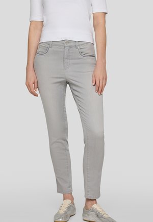 Frau trägt hellgraue Slim-Fit-Jeans, weißes Top mit halblangen Ärmeln und silberne Sneakers, steht vor einem schlichten hellgrauen Hintergrund.