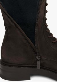 Brązowy zamszowy botek sięgający kostki z sznurowanym przodem, bocznym zamkiem błyskawicznym oraz solidną ciemną podeszwą. Wnętrze ma teksturowane wykończenie dla większego komfortu.