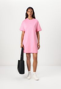 Roze katoenen T-shirt-jurk met korte mouwen, een ronde halslijn en een rechte pasvorm, gecombineerd met witte sneakers en een zwarte schoudertas.