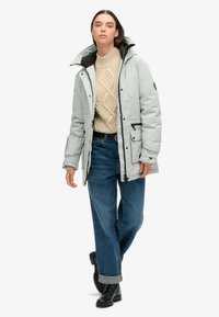 Jasnoszara puchowa kurtka z kapturem, z czarnymi akcentami i kieszeniami, noszona na kremowym swetrze w sploty i niebieskich dżinsach.