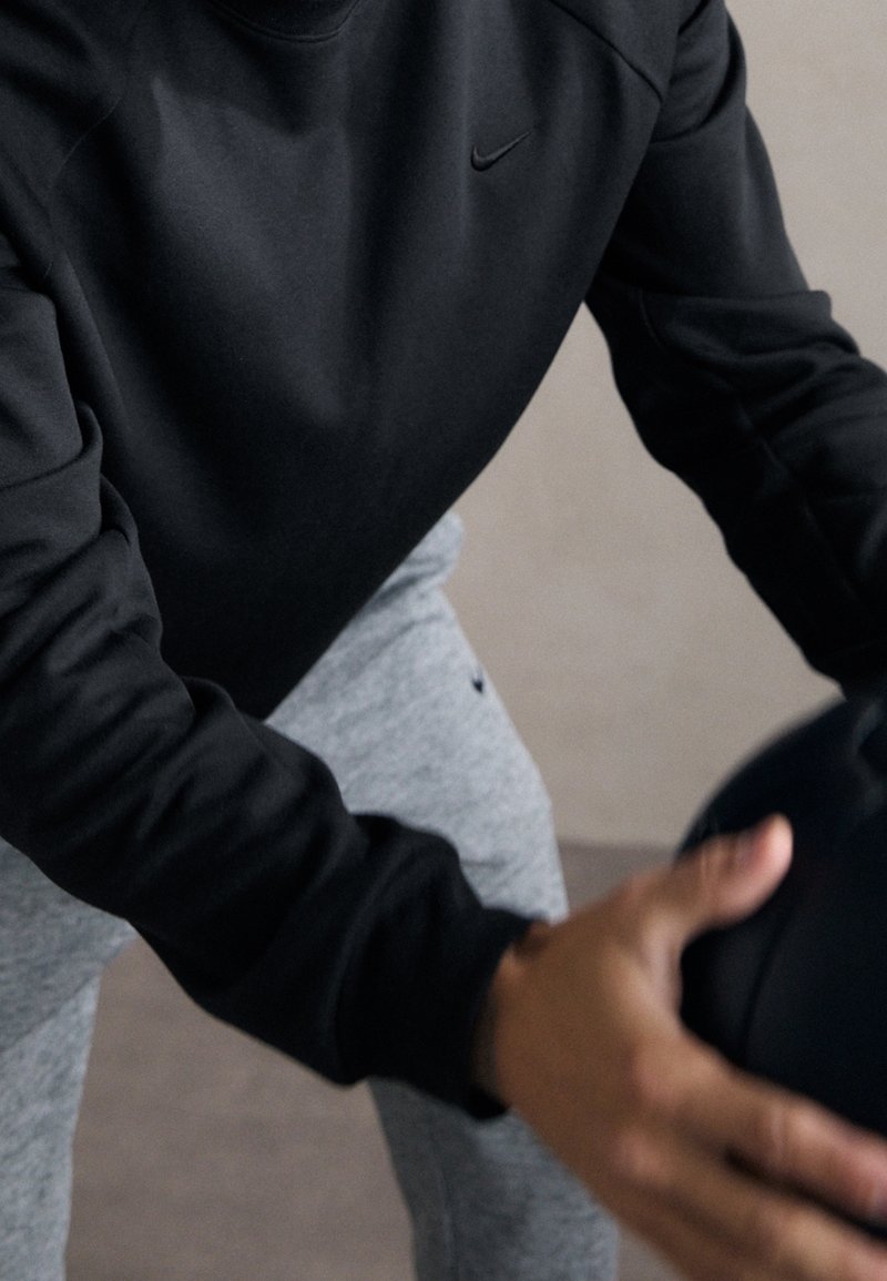 Personne portant une chemise noire à manches longues et un pantalon gris tient un ballon médicinal noir, se préparant à faire de l’exercice à l’intérieur.