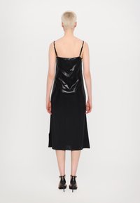 Schwarzes, ärmelloses Kleid aus glänzendem Material, midi-lange, mit dünnen Trägern und Faltenwurf-Design. Kombiniert mit schwarzen Absatzsandalen.