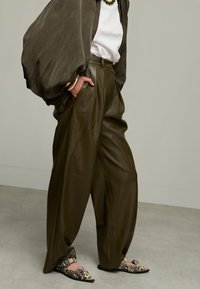 Persoon gekleed in overgrote olijfgroene leren broek, witte top, donkere losse jas, gouden armbanden, groene ketting en gedessineerde platte schoenen die op een grijze vloer staat.