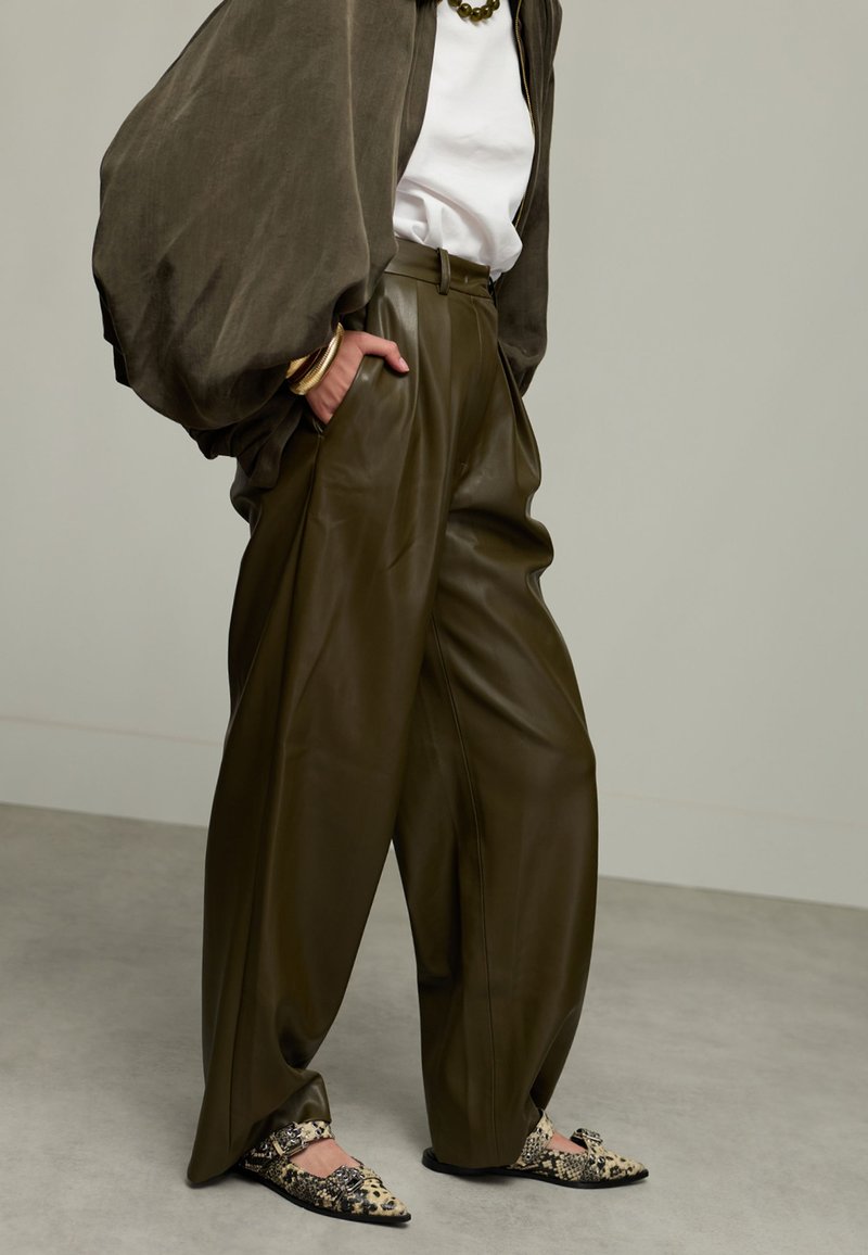 Persoon gekleed in overgrote olijfgroene leren broek, witte top, donkere losse jas, gouden armbanden, groene ketting en gedessineerde platte schoenen die op een grijze vloer staat.