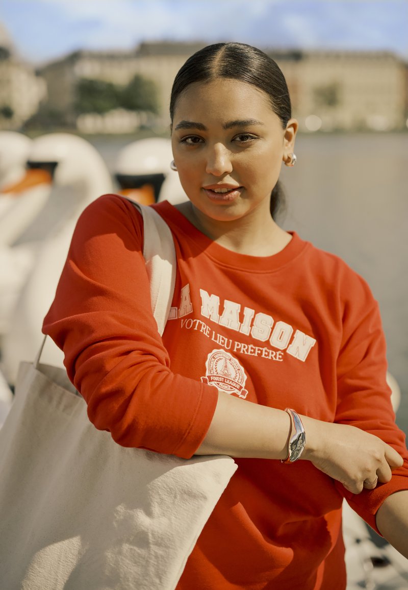 Un sweatshirt rouge avec du texte blanc et un logo, associé à un sac fourre-tout beige. La scène présente des bateaux-cygnes en arrière-plan.