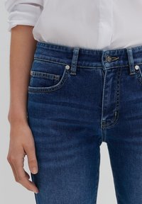 Kvinna i högmidjade blå denimjeans och en vit långärmad skjorta, med handen avslappnat vid sidan.