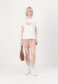 T-shirt blanc en coton avec un design abstrait corail, associé à un short rayé rose clair. Le modèle tient un sac à main marron et porte des baskets blanches.