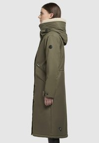 Lange olijfgroene parka met een zachte gevoerde capuchon, zijritsen en een logo-applicatie op de mouw. Strak, waterafstotend materiaal.