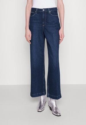 Kobieta ubrana w wysokie, szerokie dżinsy w ciemnoniebieskim kolorze, zestawione z metalicznymi srebrnymi botkami na kostkę i białą, włożoną do środka bluzką.