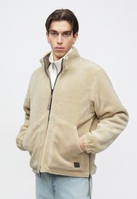 Beige fleecejakke med høj krave, lynlåslukning og to forlommer. Har elastiske manchetter og et lille brandmærke på kanten.