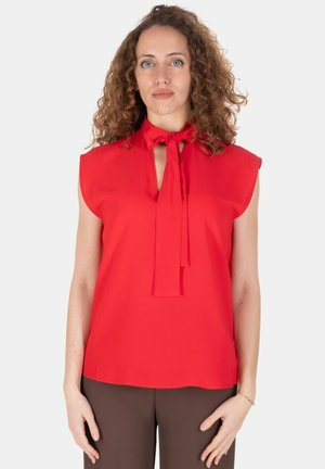 Donna con capelli ricci che indossa una camicetta rossa senza maniche con un fiocco annodato al collo e pantaloni marroni, in piedi davanti a uno sfondo bianco.