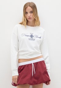 Joven con cabello rubio que lleva un sudadera blanca con el texto "New York" y una falda mini roja con cordón blanco.