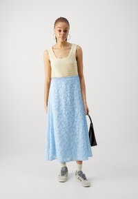 Krém színű kötött crop top, hullámos szegéllyel, világoskék maxi szoknyával párosítva, amely virágos hímzéssel díszített. A modell egy fekete táska a kezében tart.