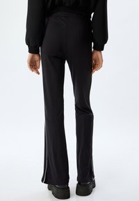 Zwarte flared pants met een gladde textuur en een hoge taille. Heeft een slit op de buitennaad en een elastische tailleband voor comfort. Zwarte schoenen zichtbaar.