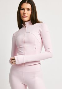 Lys rosa treningsjakke med glidelås laget av elastisk stoff. Har høy krage, figursydd design og hull for tomlene i ermene for ekstra komfort.