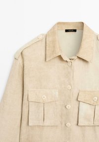 Beige Leinenhemd mit Kragen, zwei Fronttaschen und Knopfverschlüssen. Verfügt über Schulterknöpfe und eine strukturierte Oberfläche.