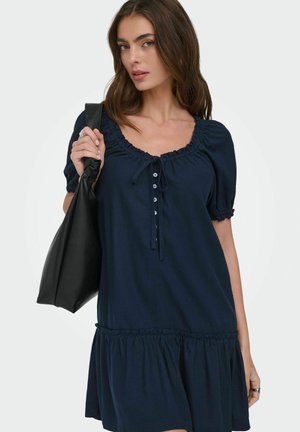 Kvinde iført mørkeblå kjole med korte ærmer og knapper, bærende en stor sort skuldertaske mod en ensfarvet lys baggrund.