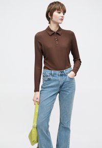 Smeđa rebrasta polo majica dugih rukava s dugmićima, uparena sa svijetloplavim denim trapericama širokih nogavica. Opremljena torbom u limun-zelene boje.