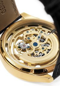 Gullfarget skjelettur med gjennomsiktig bakdeksel som avslører intrikate urverk med blå aksenter. Svart lærrem med teksturert overflate.