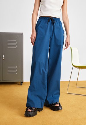 Personne portant un pantalon large bleu avec un cordon noir, des sandales plateformes noires et un haut blanc sans manches, debout à l'intérieur.