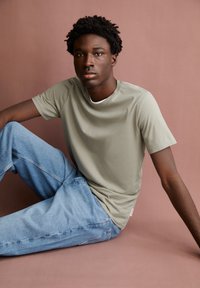 Light olive green t-shirt with a crew neck, paired with light blue jeans. Fabric appears smooth and casual, seated against a muted pink background.