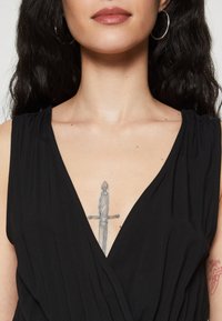 Μαύρο αμάνικο φόρεμα τύπου wrap με βαθύ V-ντεκολτέ και μαζεμένα σχέδια στους ώμους. Ορατό τατουάζ ενός σπαθιού στην περιοχή του στήθους.