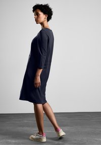 Robe bleu marine midi avec des manches trois-quarts, présentant une coupe simple et un petit détail avec un bouton à l'arrière, portée avec des baskets beiges et roses.