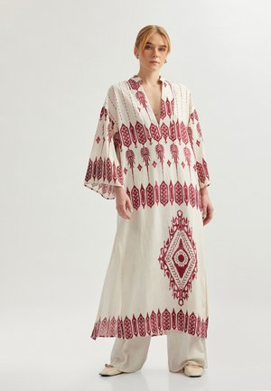Weißes Kleid mit roten gestickten geometrischen Mustern, V-Ausschnitt, langen Ärmeln und fließendem Design. Kombiniert mit hellbeigen Hosen und Schuhen.