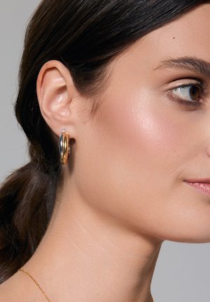 Złote i srebrne kolczyki w kształcie obręczy o gładkim, błyszczącym wykończeniu, przedstawiające splątane okrągłe kształty na neutralnym tle.