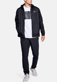Chaqueta ligera negra con capucha, presenta una impresión del logotipo gris en el pecho, combinada con pantalones negros y zapatillas deportivas de color gris claro.