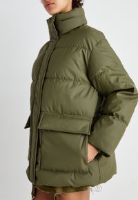 Chaqueta acolchada verde oliva con cuello alto, grandes bolsillos y diseño acolchado. Textura suave con un acabado ligeramente brillante.