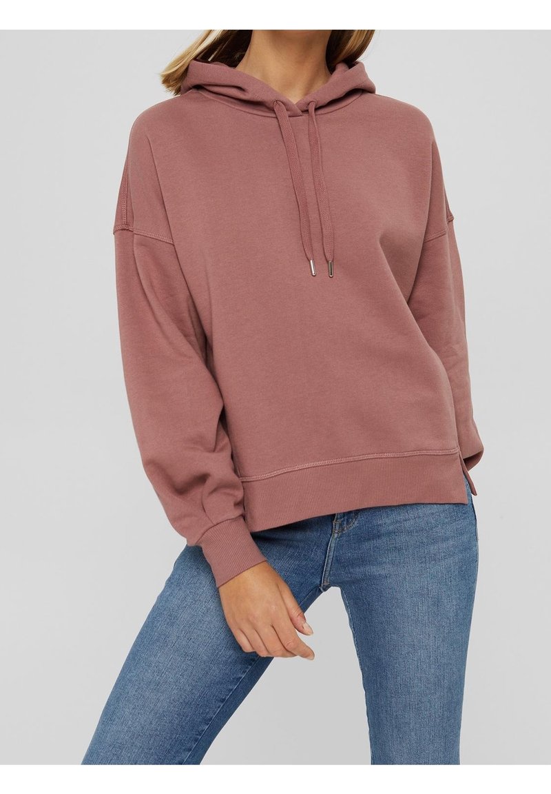 Femme portant un sweat à capuche rose poudré avec cordons et manches retroussées, associé à un jean bleu sur un fond gris clair uni.