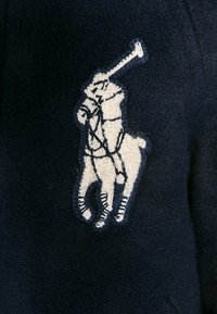 Mörkblå fleecejacka med en broderad polospelarelogotyp i krämfärg, med detaljerad sömnad och en texturerad yta.