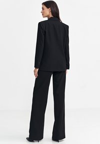 Czarny garniturowy żakiet i spodnie o szerokich nogawkach. Gładki materiał o sztywnej strukturze, z kołnierzem w kształcie notcha i guzikowymi mankietami.
