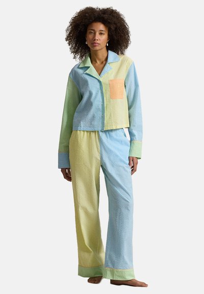 Femme aux cheveux bouclés portant un ensemble de pyjama aux couleurs pastel avec des tons bleus, verts et jaunes, debout pieds nus sur un fond blanc.