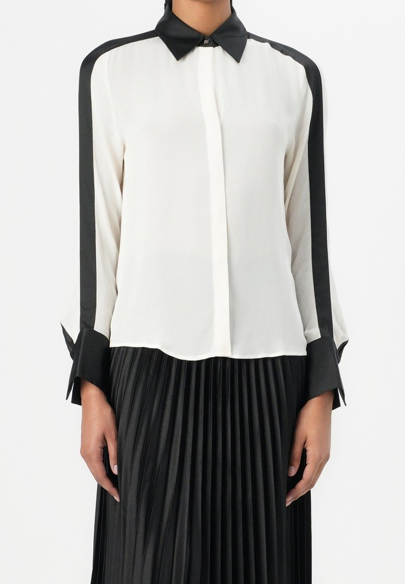 Femme portant un chemisier blanc à manches longues avec col noir, poignets noirs et rayures noires sur les manches, associé à une jupe plissée noire.