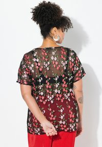Camicia nera trasparente con ricamo floreale in rosso e bianco, maniche corte, vestibilità comoda, indossata sopra un pantalone rosso con texture.